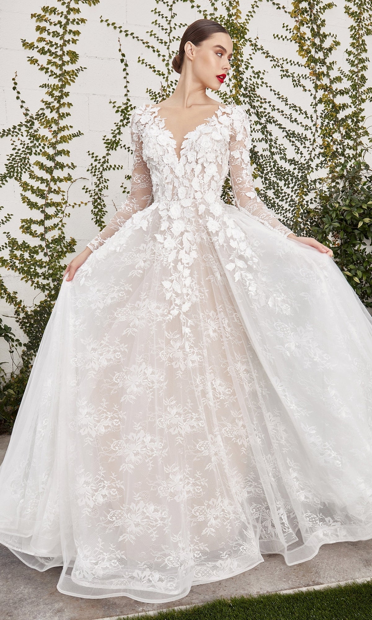 A woman stands outdoors in front of a white wall with climbing greenery, holding out the skirt of the Long Sleeve White Lace Bridal Gown: LadivineA1067W, featuring floral appliqués.